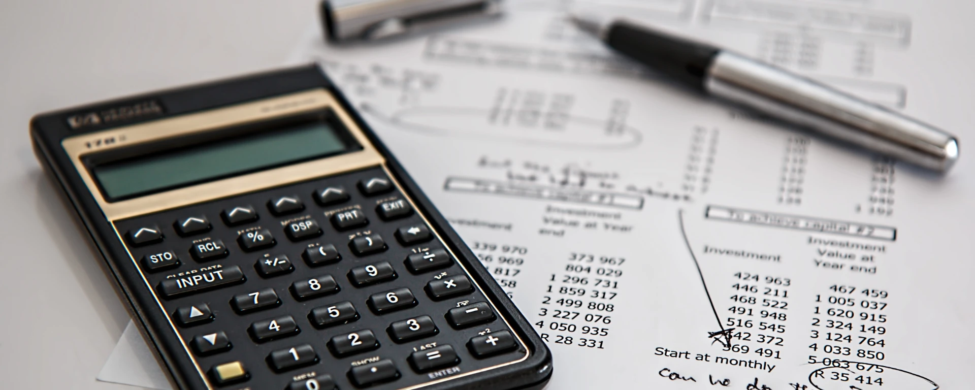 Photograph showing a black scientific calculator placed on top of printed financial documents with handwritten notes and a silver pen nearby. Documents contain numerical data, tables, and annotations related to investment values and start dates, indicating financial analysis or budgeting activity.