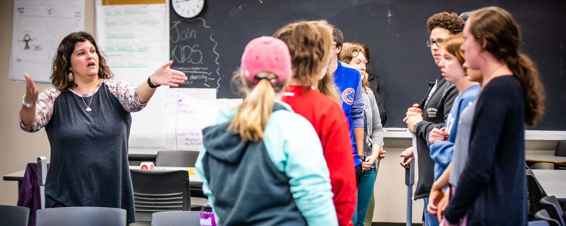 A teacher in a dark shirt gestures while speaking to a group of students standing in a classroom with a blackboard and clock visible in the background at Northwest Missouri State University
