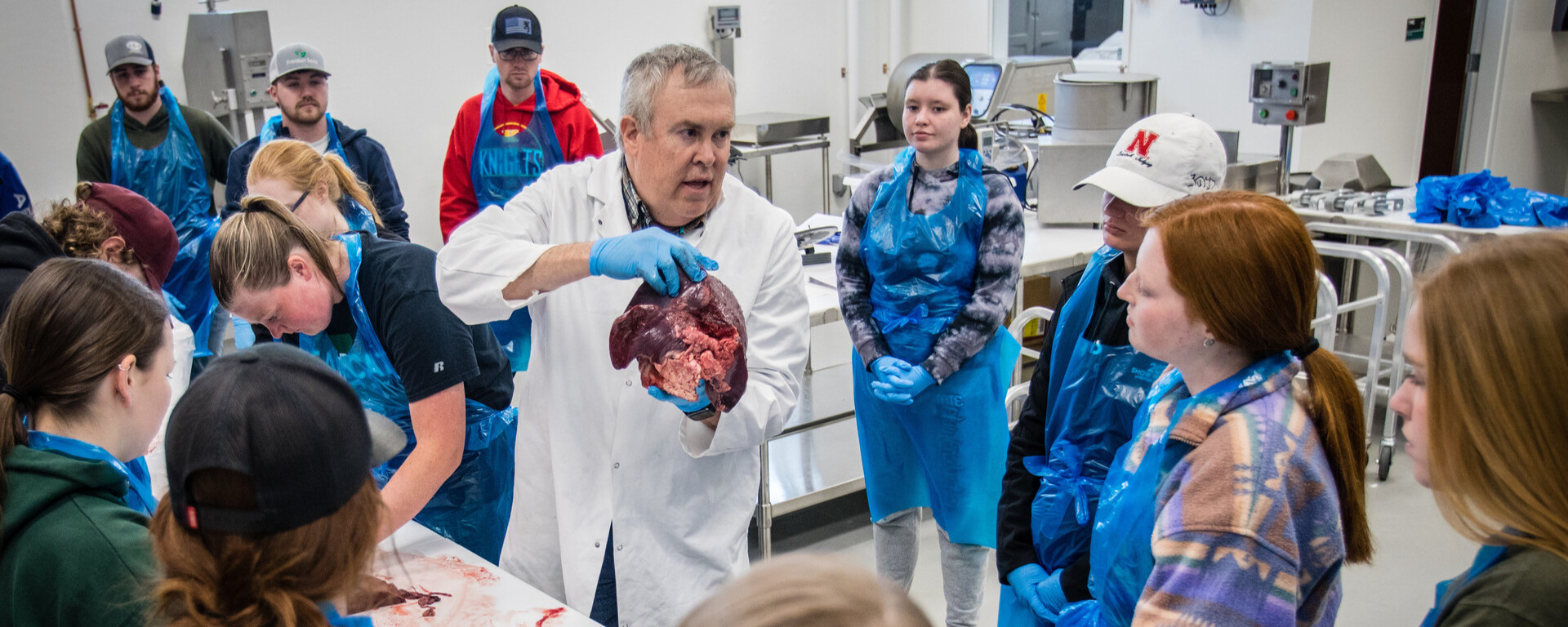 A man in a white lab coat holds and demonstrates an animal organ to a group of students wearing blue aprons in a laboratory setting at Northwest Missouri State University
