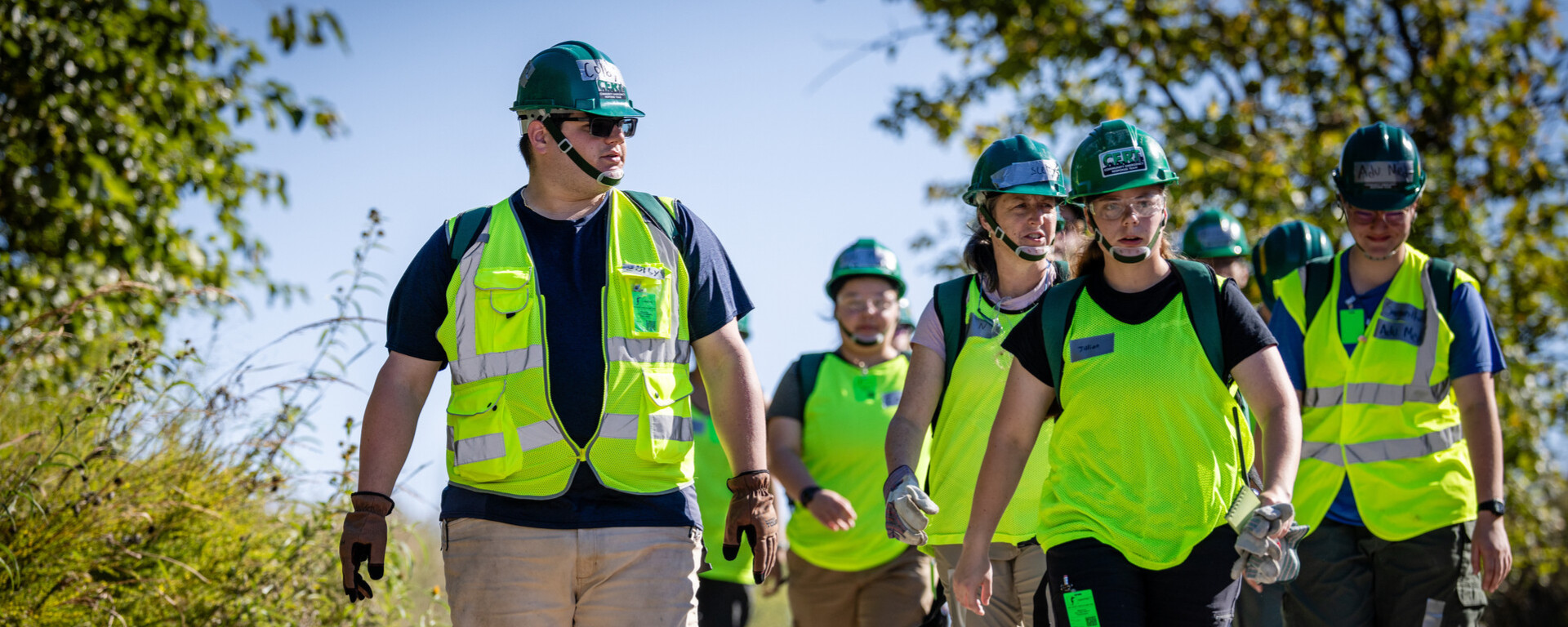 A group of workers wearing bright green safety vests and hard hats walking together on a gravel path lined with trees and vegetation at Northwest Missouri State University