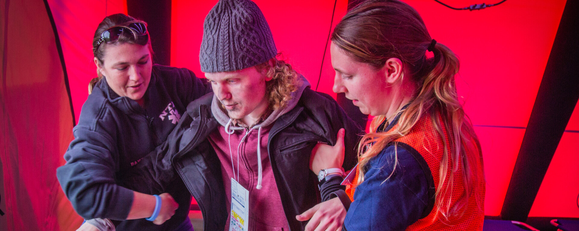 Three young people stand together in a tent or enclosed space illuminated with bright red and purple lighting. The person on the left wears a dark jacket and jeans, the middle person wears a gray beanie, pink hoodie, and dark jacket with a credential badge, and the person on the right wears an orange and blue jacket with dark pants. They appear to be engaged in conversation or activity at Northwest Missouri State University.