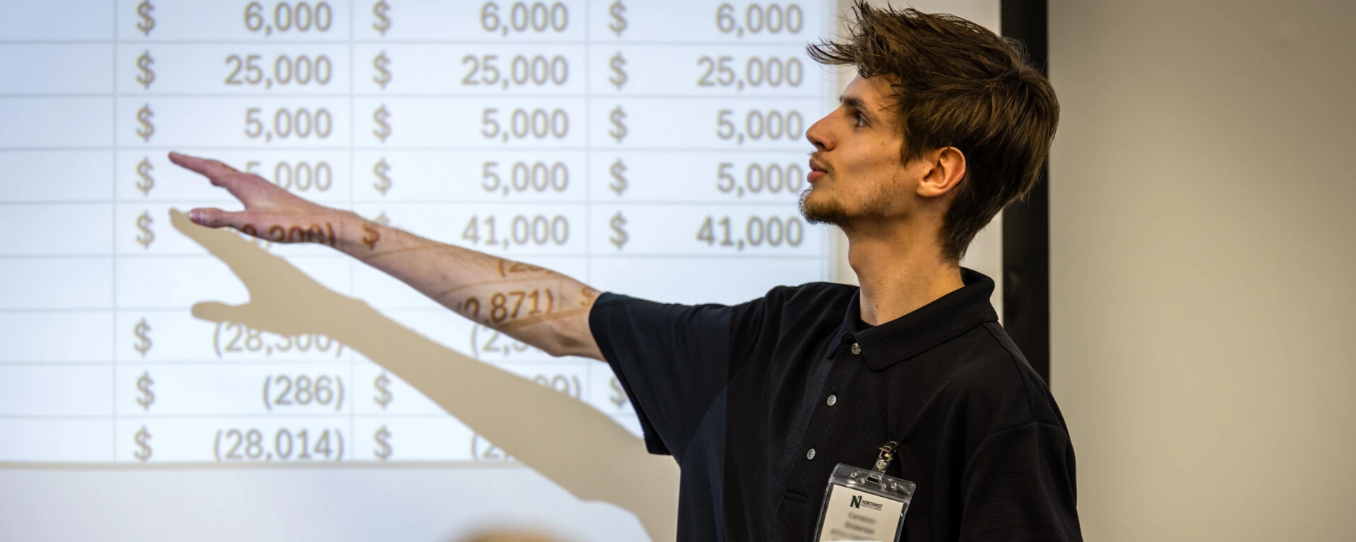A man wearing a black polo shirt and ID badge gestures toward a projected financial spreadsheet showing dollar amounts and calculations while presenting to an audience member visible in the foreground at Northwest Missouri State University