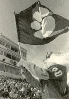 Bobby waves a Bearcat flag during a Football game.  Bobby wears overalls over his classic 00 jersey.