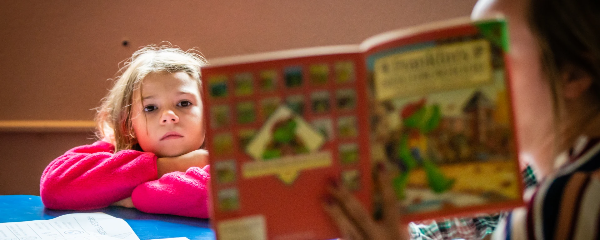 A young child sits at a table with her head laid atop her crossed arms, intently focused as the teacher reads the class a book.