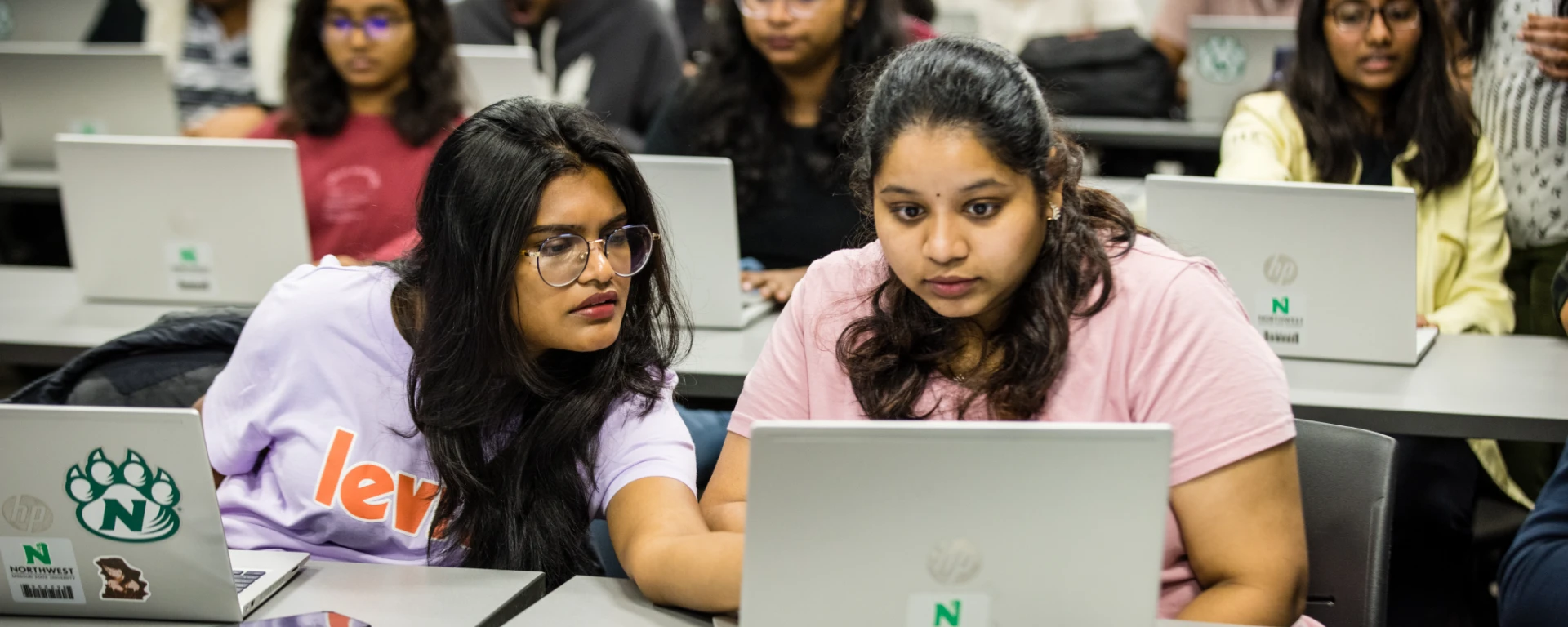 Two female students huddle around a laptop to problem-solve during their Object-Oriented Systems class