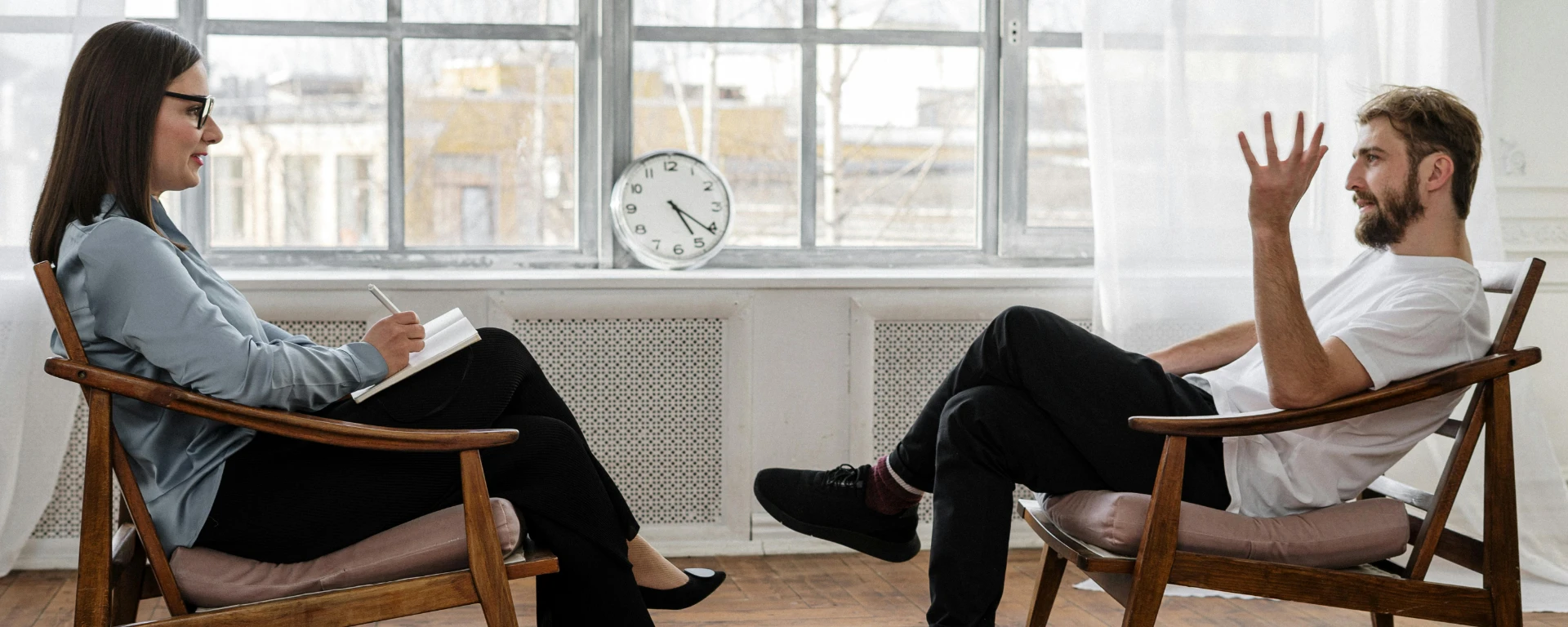 A female therapist and male patient sit face-to-face in chairs during a therapy session. The therapist holds a clipboard and pen, suggesting Cognitive Behavioral Therapy.