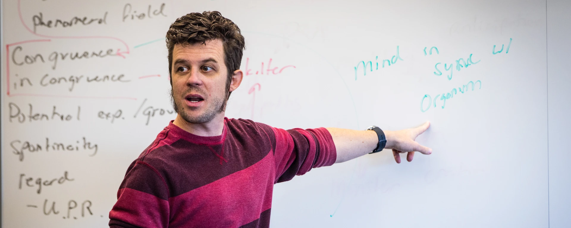 A male professor points at a whiteboard with the words 'school written'mind in sync with organism' on it during a Psychology of Personality class.