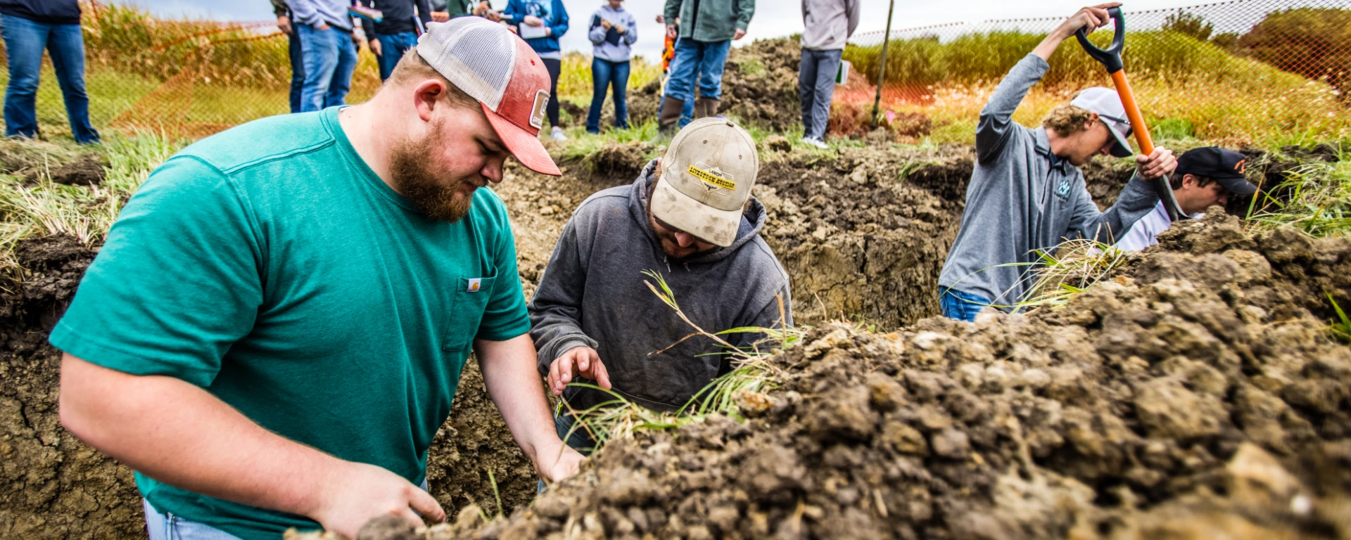 Four students stand in a deep dirt trench with shovels so they can analyze soil conditions for crop growth.
