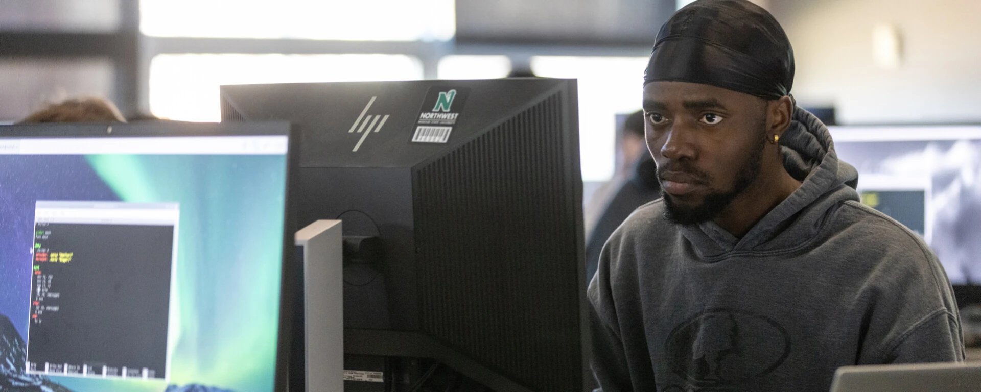 A man wearing a black headband and dark hoodie sits at a computer workstation with multiple monitors, including a black desktop tower, while working on a laptop at Northwest Missouri State University