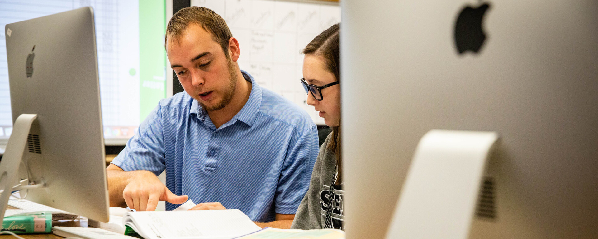 A male instructor in a blue polo shirt and a female student wearing glasses work together at a wooden desk, looking at documents and an open textbook while seated in front of Apple computer monitors in a classroom or lab setting at Northwest Missouri State University