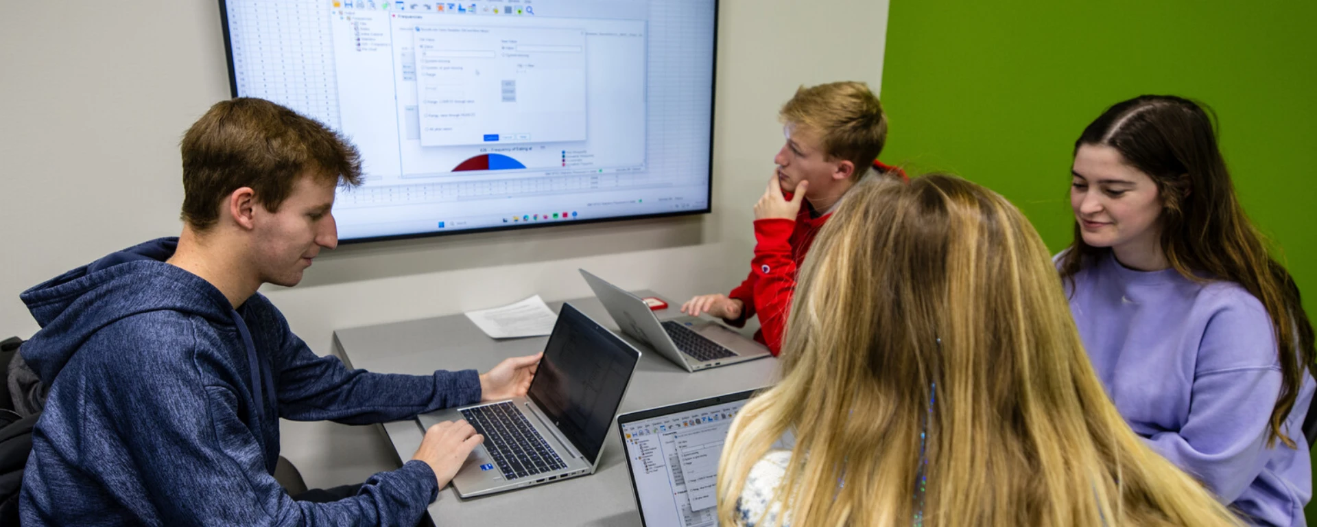 A group of five students gathered around a table with laptops and tablets, looking at a large display screen showing a software interface, in a modern room with a bright green wall at Northwest Missouri State University.