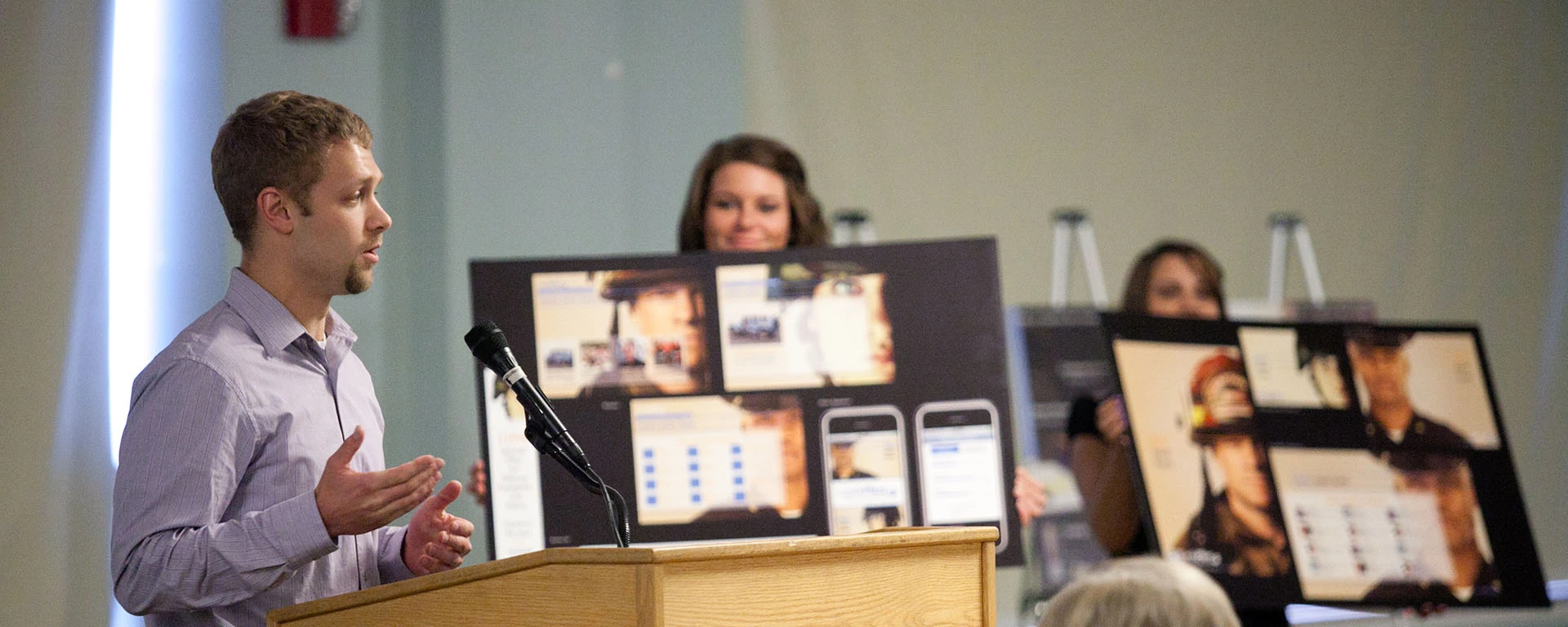 A male student stands at a podium during his team's Knacktive presentation. Visual aids of new branding and marketing ideas are in the background. 