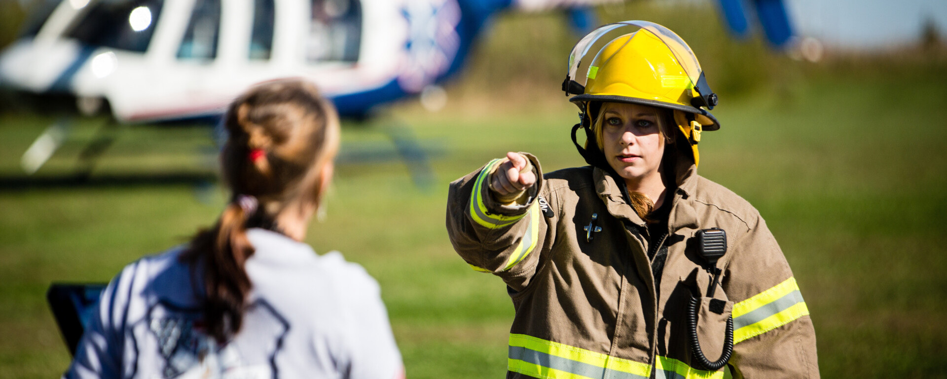 Firefighter in yellow helmet and tan gear with reflective stripes pointing while communicating with another person in front of a white and blue helicopter on a grassy field at Northwest Missouri State University