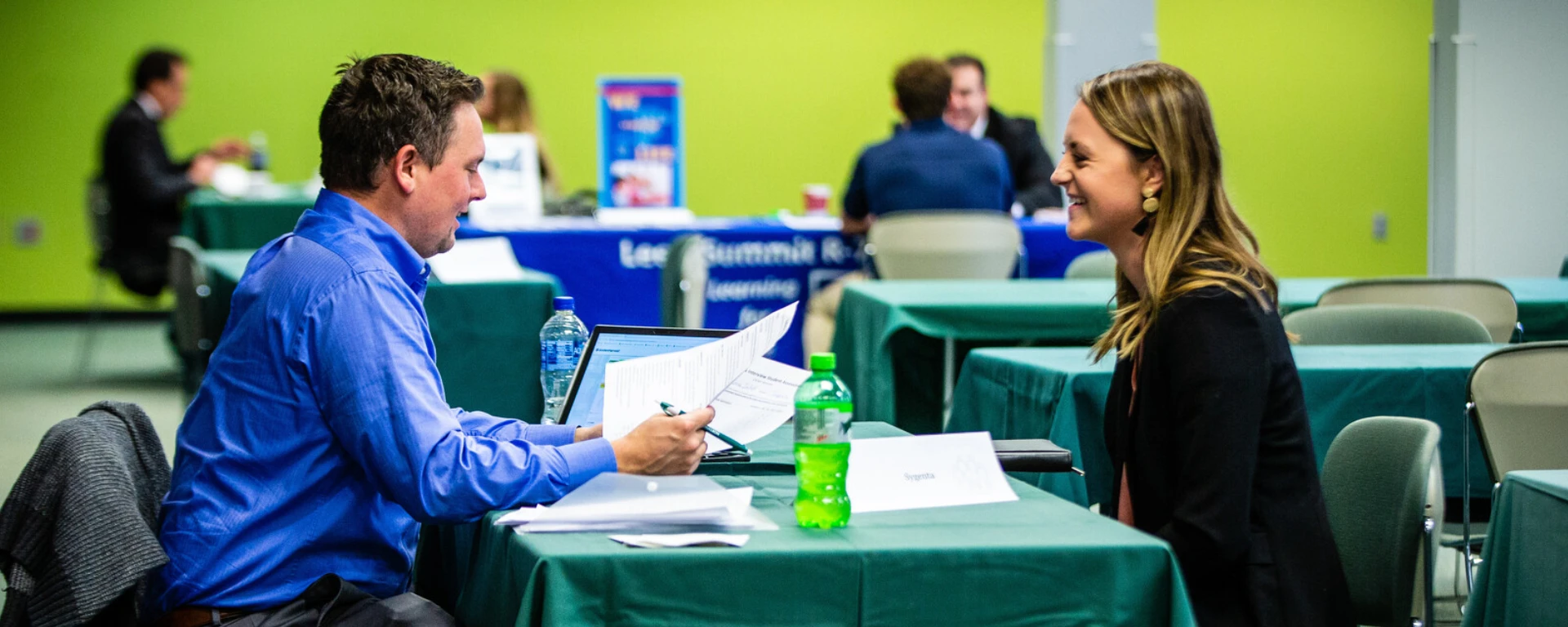 A man in a blue jacket and a woman in a black jacket conducting an interview or conversation at a green table in a bright room with lime green walls at Northwest Missouri State University