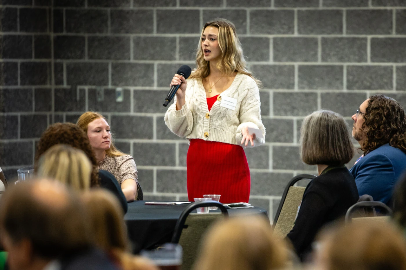 A woman with blonde hair wearing glasses, a cream cardigan, and red skirt stands holding a microphone while speaking to an audience seated in front of her against a brick wall backdrop at Northwest Missouri State University