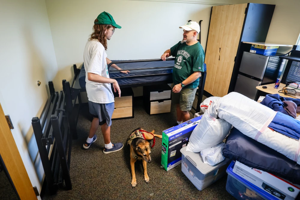 A dad helps a student organize their room during Move In Day. A service dog wearing a vest is laying on the floor nearby.